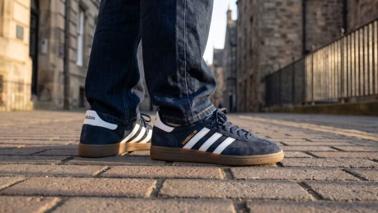 Navy blue Adidas Handball Spezial suede sneakers with white stripes and gum soles worn on a cobblestone street