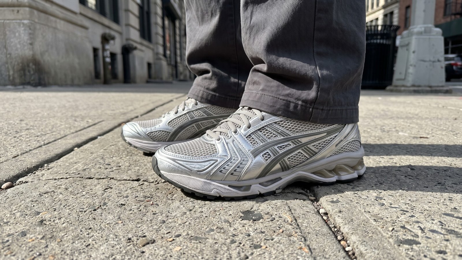 Silver ASICS Gel-Kayano 14 sneakers worn with grey trousers on a city sidewalk
