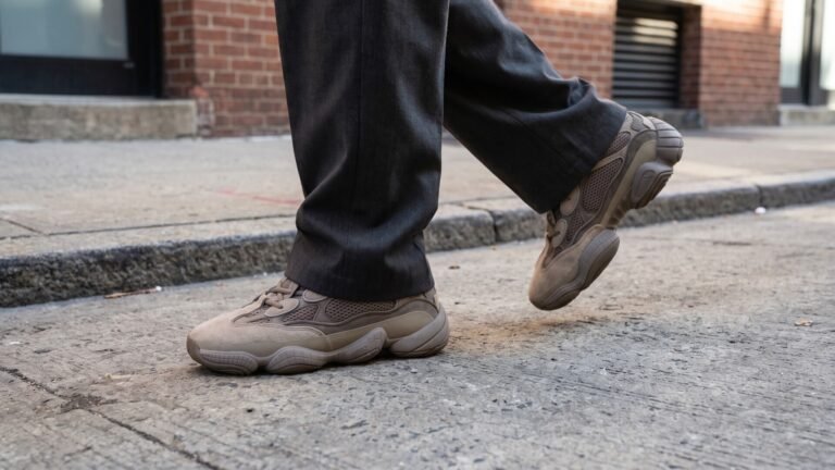 Close-up of a person walking on a concrete sidewalk wearing chunky taupe dad sneakers and dark grey trousers