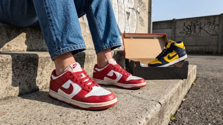 Person wearing red and white Nike Dunk Low sneakers next to a navy and yellow Dunk High on concrete steps
