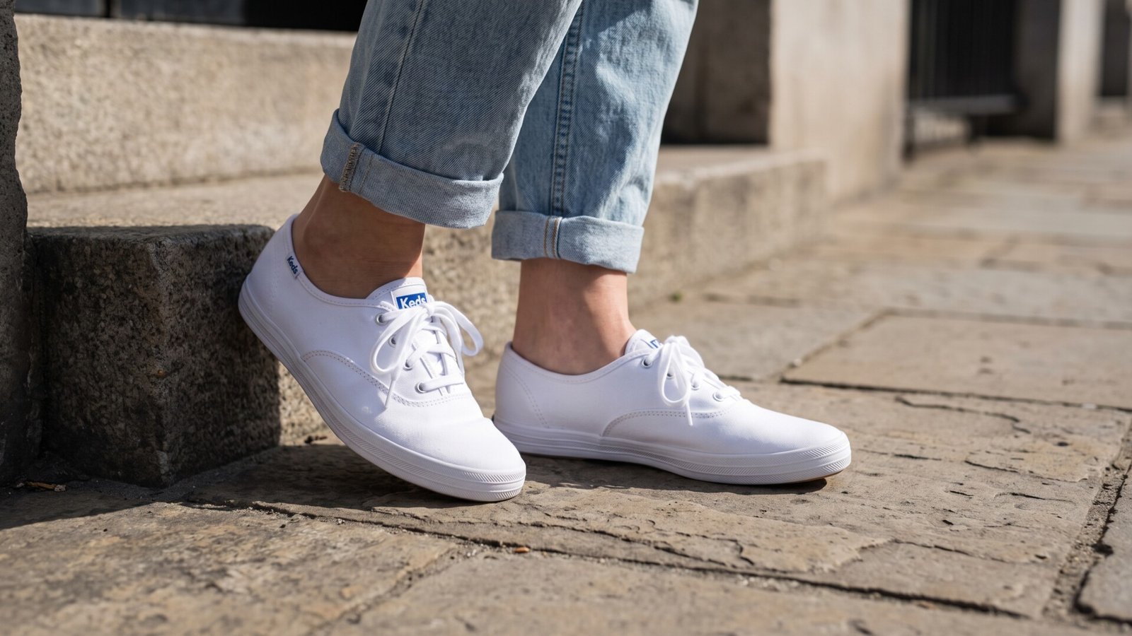 White low-profile Keds canvas sneakers worn with cuffed light-wash blue jeans on stone steps