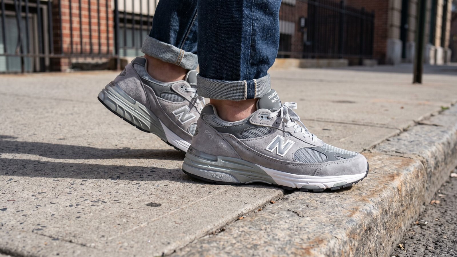 Grey New Balance 993 suede sneakers paired with cuffed blue jeans on a city sidewalk curb