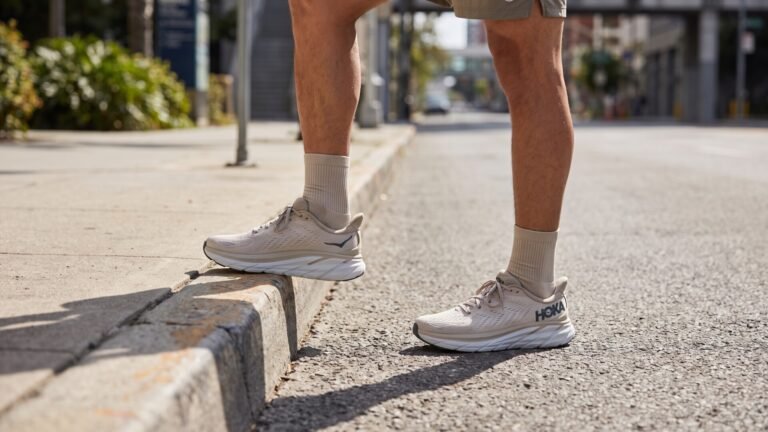 Runner wearing neutral Hoka Clifton 9 shoes paused at a sidewalk curb on a city street