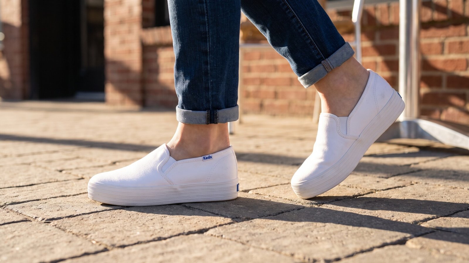White Keds Double Decker slip-on canvas sneakers worn with cuffed blue jeans on a paved outdoor walkway