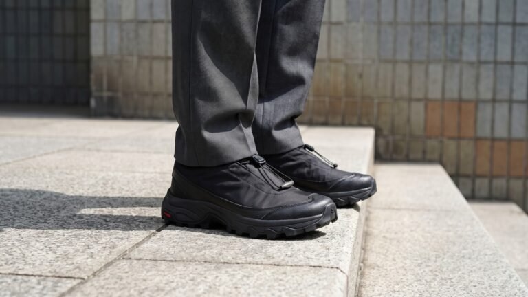 Black MM6 Maison Margiela x Salomon concept sneakers styled with grey trousers on urban stone steps