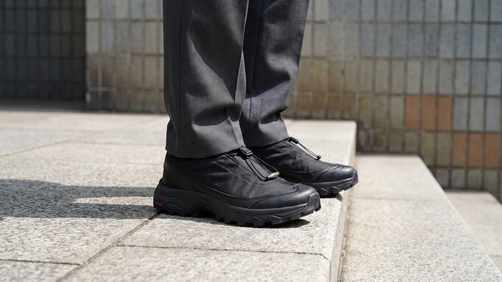 Black MM6 Maison Margiela x Salomon concept sneakers styled with grey trousers on urban stone steps