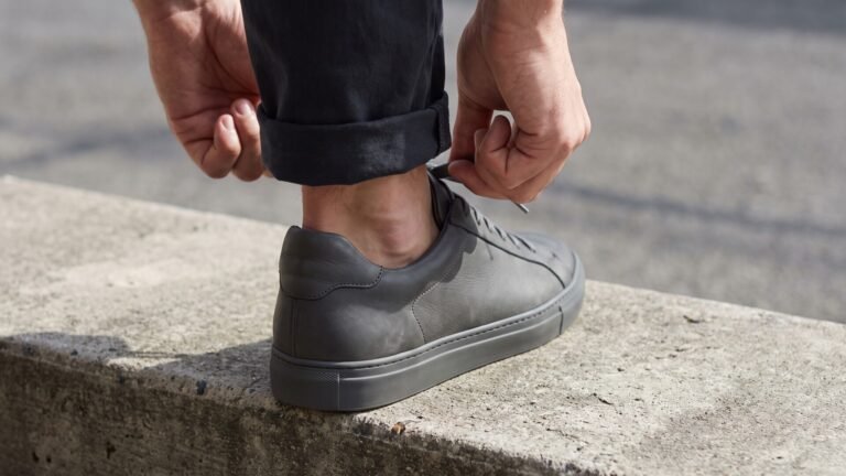 Close-up of a person tying the laces of a sleek grey leather sneaker with a snug heel fit on a concrete curb