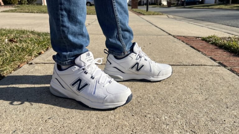 White and navy New Balance 608v5 cross-training sneakers worn with blue jeans on a concrete sidewalk