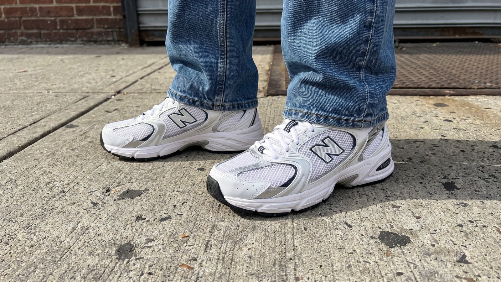 White and silver New Balance 530 sneakers paired with blue jeans on a concrete sidewalk