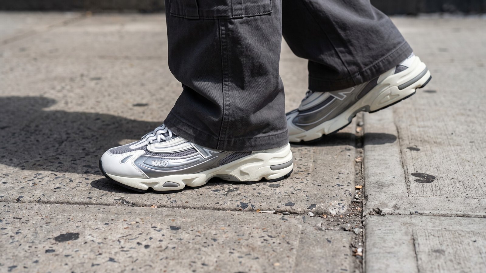 Silver and white New Balance 1000 sneakers worn with dark trousers while walking on a concrete sidewalk