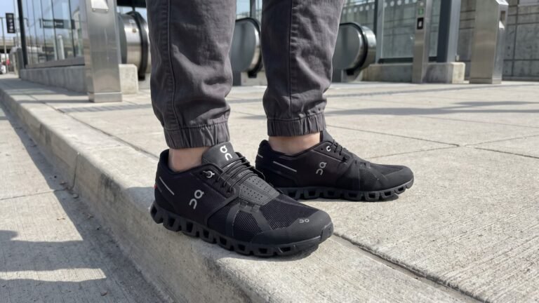 Side view of a person wearing all-black On Cloud 5 sneakers while standing on a concrete sidewalk curb
