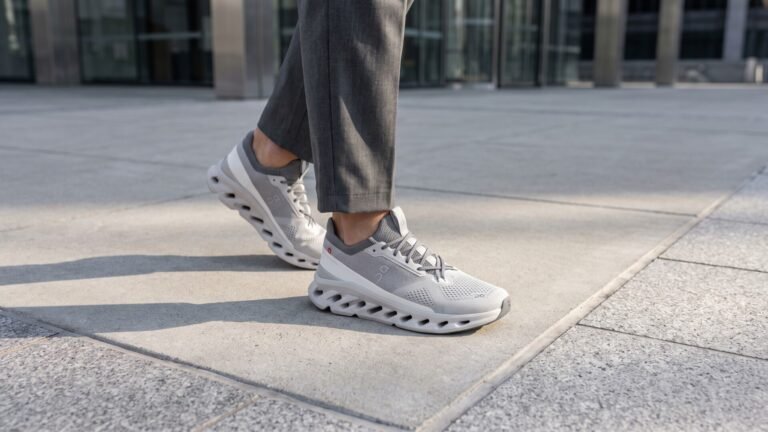Person walking on a paved city sidewalk wearing light grey On Cloudtilt sneakers and grey trousers
