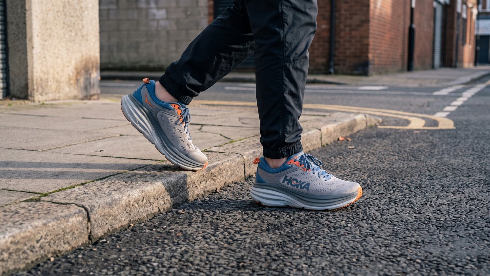 Person wearing grey and blue maximalist Hoka sneakers stepping off a sidewalk curb onto asphalt