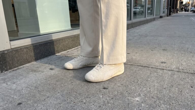 Person wearing off-white fuzzy suede Puma Speedcat sneakers and tailored cream trousers on a city sidewalk