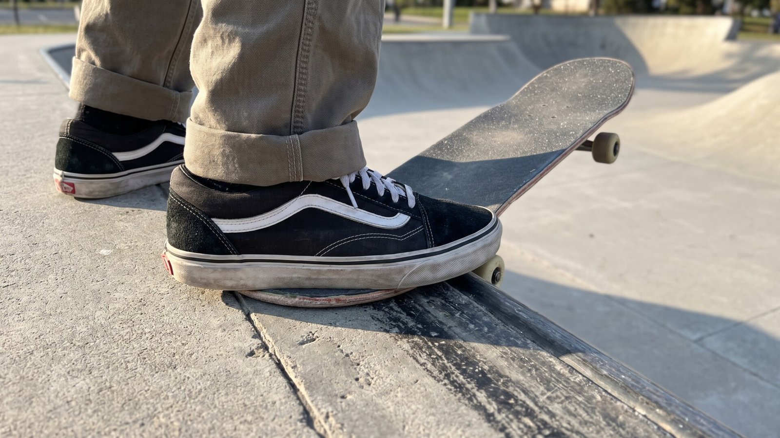 Skater wearing black Vans Old Skool sneakers balancing on a skateboard at a concrete skatepark coping