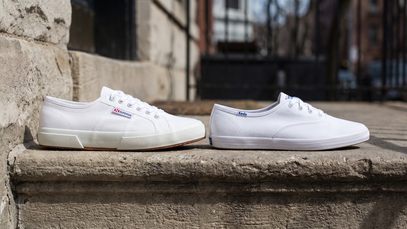 White Superga 2750 sneaker next to a white Keds Champion sneaker resting on an outdoor stone ledge