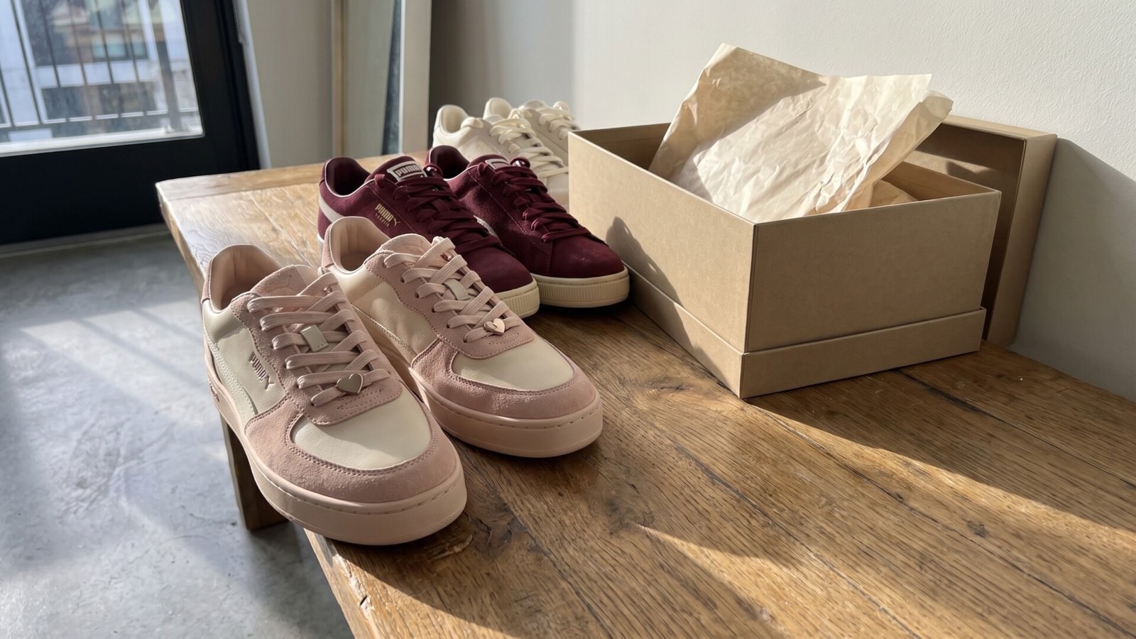 Blush pink Puma sneakers with heart charms and burgundy suede pairs displayed on a wooden bench next to a shoebox