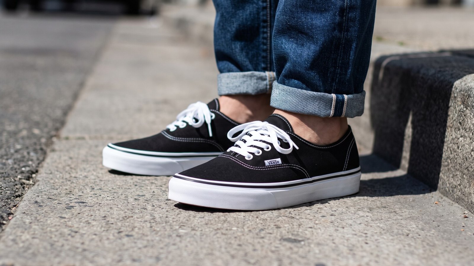 Classic black Vans Authentic canvas sneakers worn with cuffed denim jeans on a concrete sidewalk curb