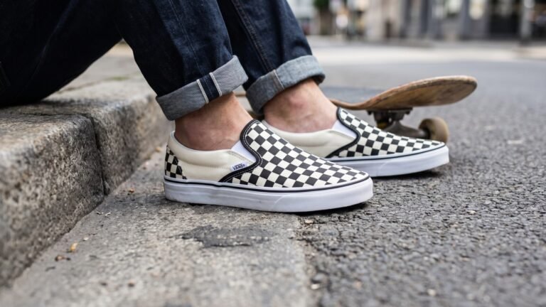 Checkerboard Vans Slip-On sneakers worn by a person sitting beside a skateboard on a city street curb